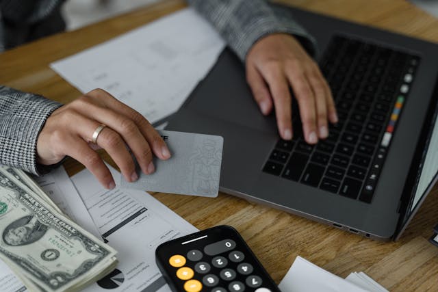Person using a laptop while holding a bank card, with money and a calculator spread out across the table